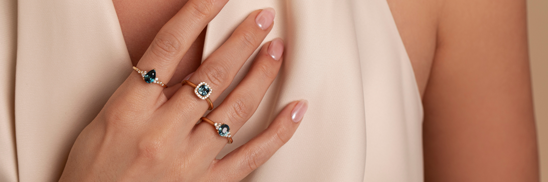 Three 18ct yellow gold teal sapphire and diamond rings worn on a model's hand, featuring pear, cushion and oval cut sapphires with different diamond settings.